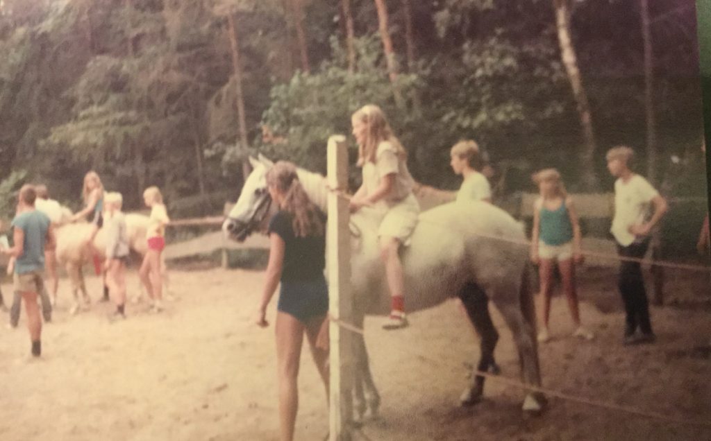 Clara riding a loan pony at pony camp around 1980, with other children and ponies in a riding arena surrounded by trees.